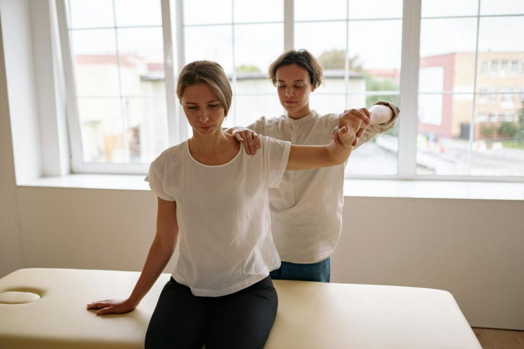 A therapist assists a woman with arm stretching during a physical therapy session in a bright, indoor setting.