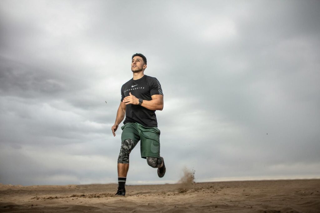 A muscular man running on sandy terrain under cloudy skies, promoting fitness and adventure.